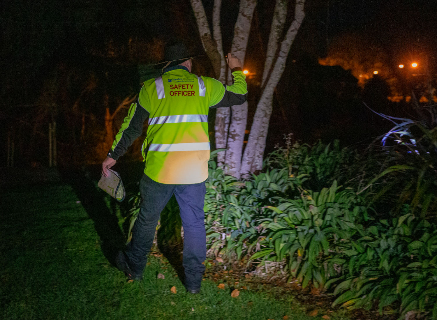A man with a yellow high visibility jacket, with City Safe written on it, is on patrol in the dark. He is shining a torch behind some trees in a park.