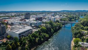 An aerial view of Hamilton City with the Waikato river on the right and a bridge in the distance. On the left bank of the river are trees with city buildings behind and stretching to the horizon.